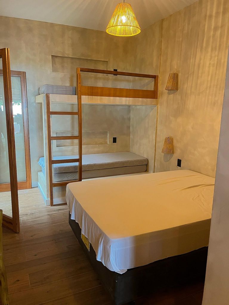 Quarto da casa com cama de casal arrumada e um beliche de madeira ao lado