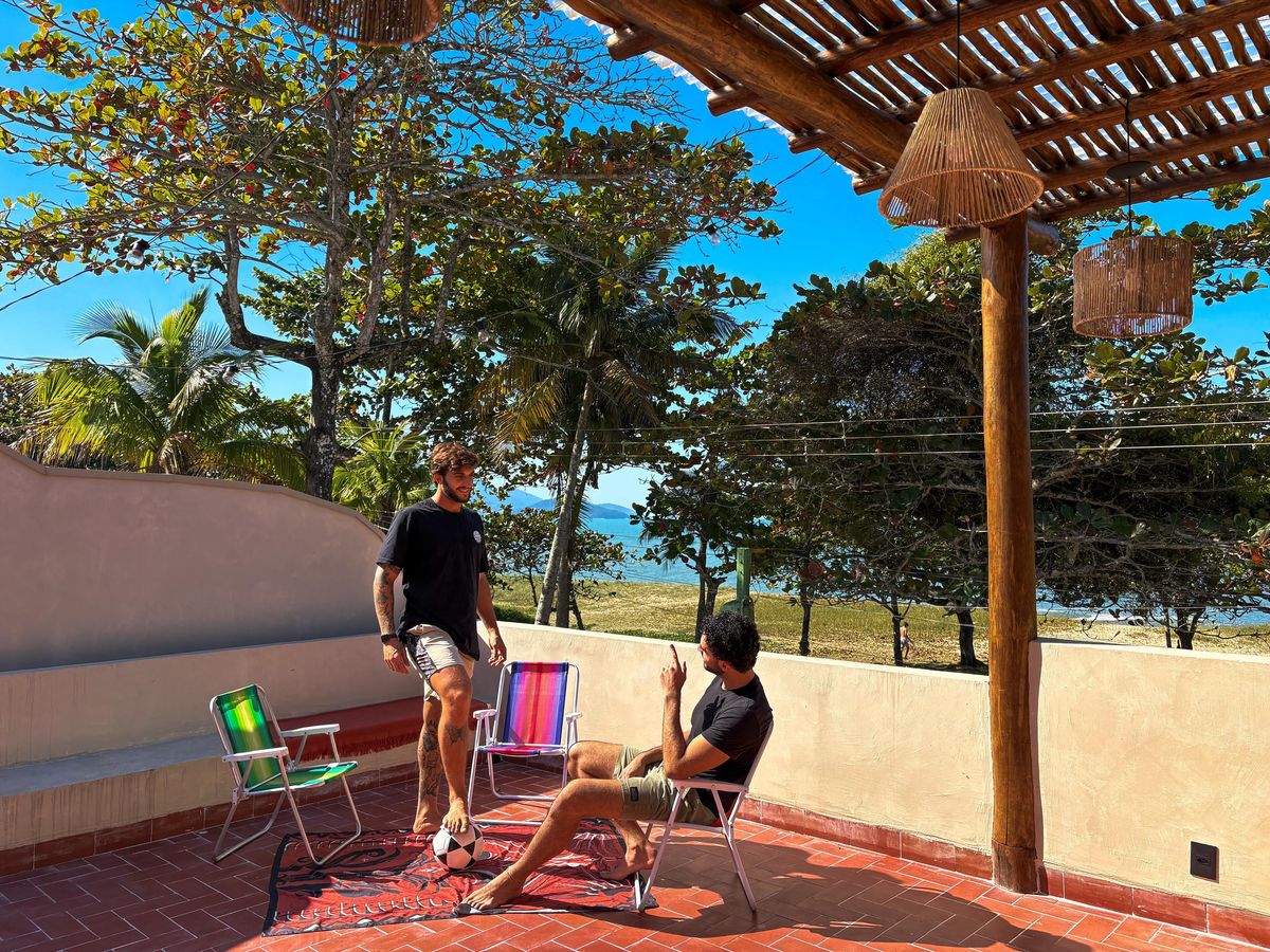 Dois hóspedes no rooftop da casa, sob a pérgola de madeira, com cadeiras de praia e o mar entre as árvores