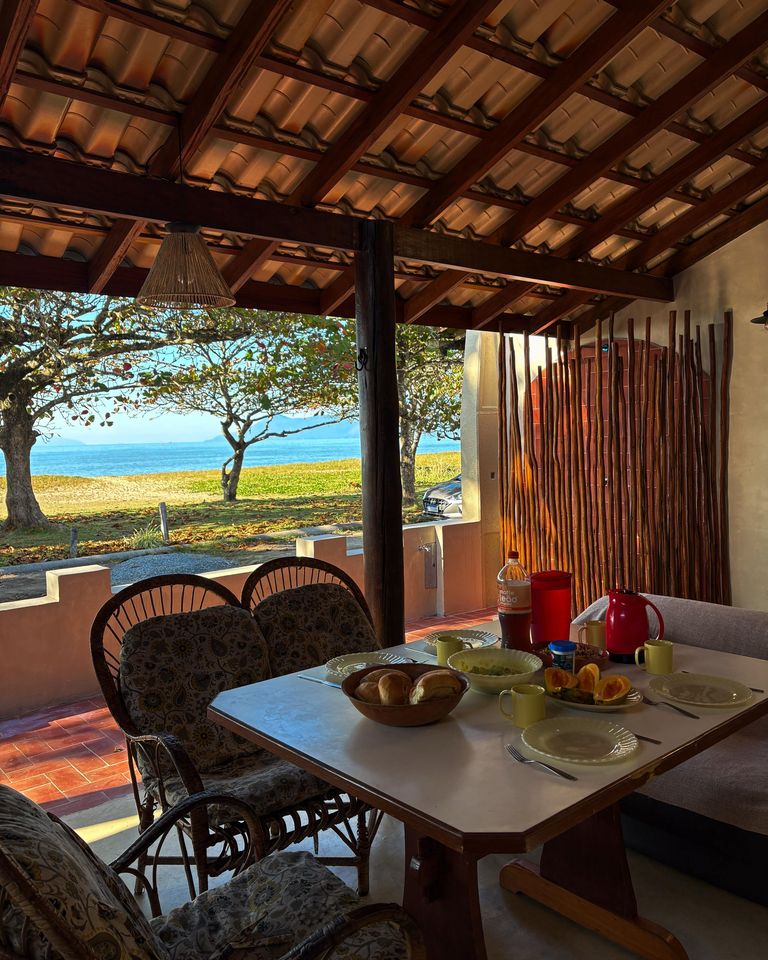 Mesa posta na varanda coberta da casa, com o mar visível entre as árvores